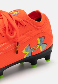 Scarpa da calcio sintetica arancione con suola nera e tacchetti verdi. Presenta una parte superiore testurizzata e un logo grigio e giallo ben visibile.