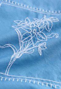 Blauwe stof met witte bloemgeborduurde motieven, gedetailleerde bloemblaadjes en bladeren, en zichtbare stiksels langs de randen.