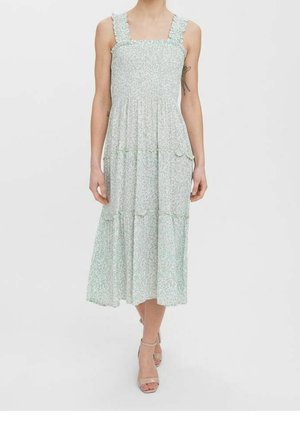 Femme portant une robe sans manches, longue jusqu'aux chevilles, bleu clair à fleurs avec des bretelles volantées et des talons beiges à bride à la cheville, debout devant un fond uni.
