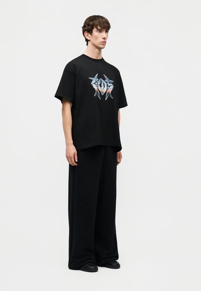 Νέος άνδρας που στέκεται φορώντας μαύρο φαρδύ παντελόνι, μαύρο t-shirt με μεταλλικό σχέδιο αστεριού και μαύρα παπούτσια σε λευκό φόντο.