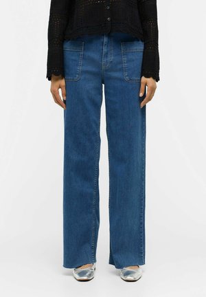 Femme portant un jean bleu à jambes larges avec de grandes poches avant, associé à un haut noir texturé à manches longues et des ballerines argentées.