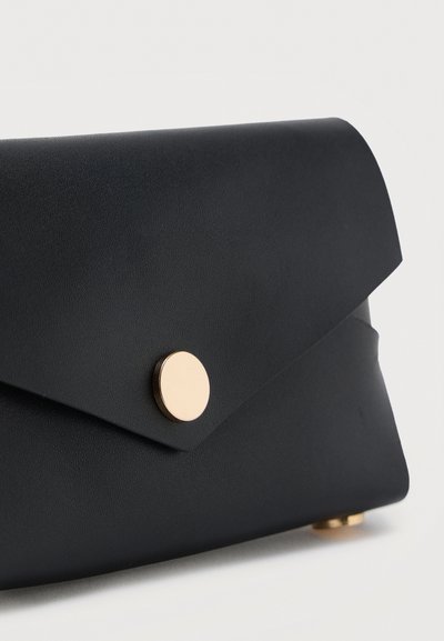 Μαύρο δερμάτινο τσαντάκι χειρός με χρυσό κυκλικό κούμπωμα και λοξή πτερύγιο, τοποθετημένο σε λευκή επιφάνεια.