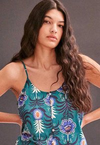 Femme aux longs cheveux ondulés et foncés, portant un haut sans manches bleu sarcelle avec des motifs floraux, debout les mains sur les hanches devant un fond uni.