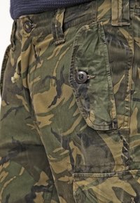 Cargo-byxor i ett grönt kamouflage-mönster. Funktioner inkluderar knäppbara sidofickor, en dragsko i midjan och en bomullsblandning.