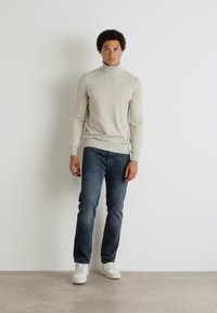Beige garbónyakú pulóver ribbelt texture-rel, sötétkék farmerrel és fehér sportcipővel párosítva. A modell egy sima háttér előtt áll.