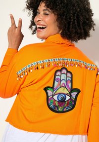 Orange kort jacka med fransdetaljer, som har en flerfärgad broderad handdesign med ett öga på baksidan.