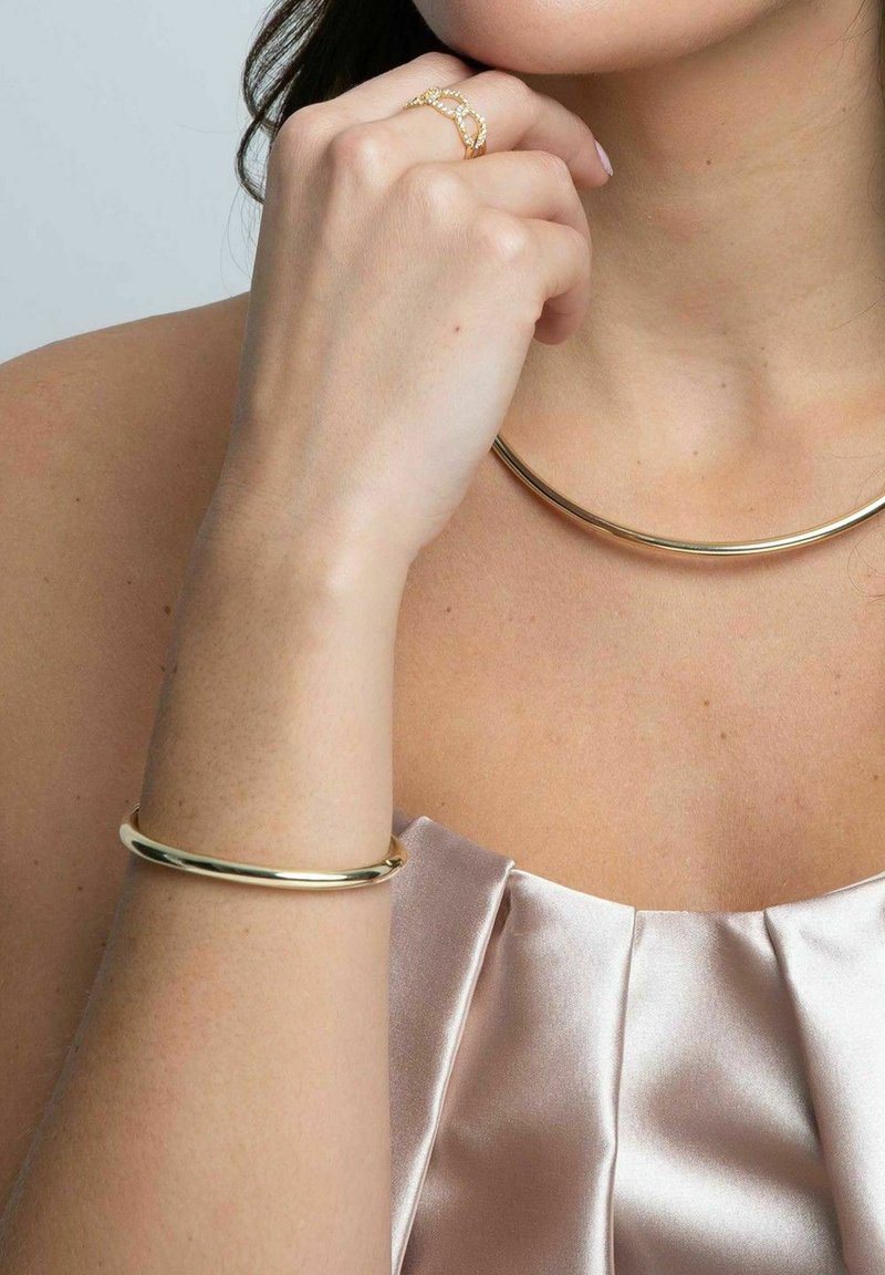 Guldkedja och armband på en person som bär en satinfärgad blus i blekrosa, med en delikat ring på handen. Släta, polerade ytor.