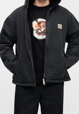 Person iført en sort lynlåstrøje over en sort t-shirt med en blæksprutte- og citrongrafik på brystet.