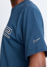 Osoba v modrém tričku Nike s malým bílým logem Nike swoosh na rukávu a částečně velkým grafickým motivem na hrudi.