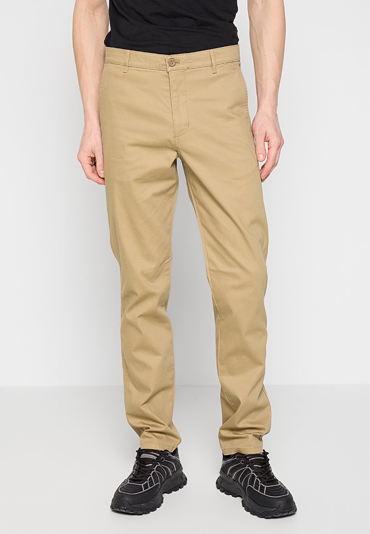 Beige katoenen broek met een rechte snit, knoopsluiting en riemlussen, gecombineerd met zwarte sneakers met gestructureerde zolen en mesh accenten.