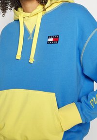 Sudadera azul y amarilla con un bolsillo delantero, que incluye capucha, cordones y el logotipo "TOMMY JEANS" en un parche rojo, blanco y azul.