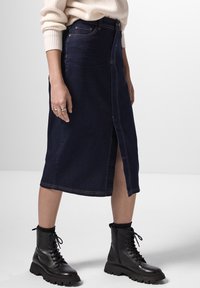 Falda midi de mezclilla azul oscura con una abertura frontal, acentuada con costuras doradas. Combinada con botas de combate negras con cordones para un look casual.