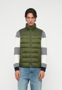 Vesta puf în verde măsliniu, cu guler înalt și închidere cu fermoar. Țesătura este netedă; mânecile au un model în dungi în gri și alb.