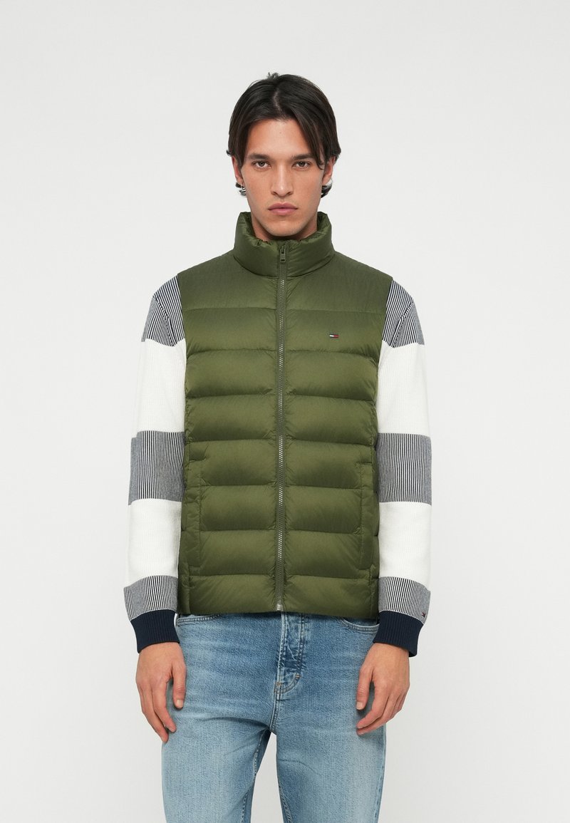 Vesta puf în verde măsliniu, cu guler înalt și închidere cu fermoar. Țesătura este netedă; mânecile au un model în dungi în gri și alb.