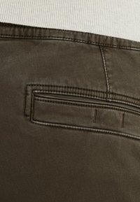 Tissu vert olive foncé avec une texture lisse, présentant une poche arrière avec des bords cousus et une ceinture droite au-dessus.