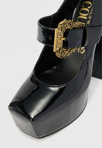 Zapatos de tacón de plataforma de charol negro con puntera redonda, correa en el tobillo y detalles ornamentales de hebilla dorada. Textura suave, tacón alto y cuadrado.