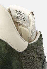 Zapatilla de ante verde con tejido texturizado y un acento de cuero color crema. La etiqueta interior presenta una cita motivacional.
