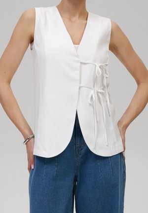 Ärmelloses weißes Wickeltop mit zwei seitlichen Bändern, getragen zu hoch taillierten blauen Jeans, silbernes Armband am linken Handgelenk.