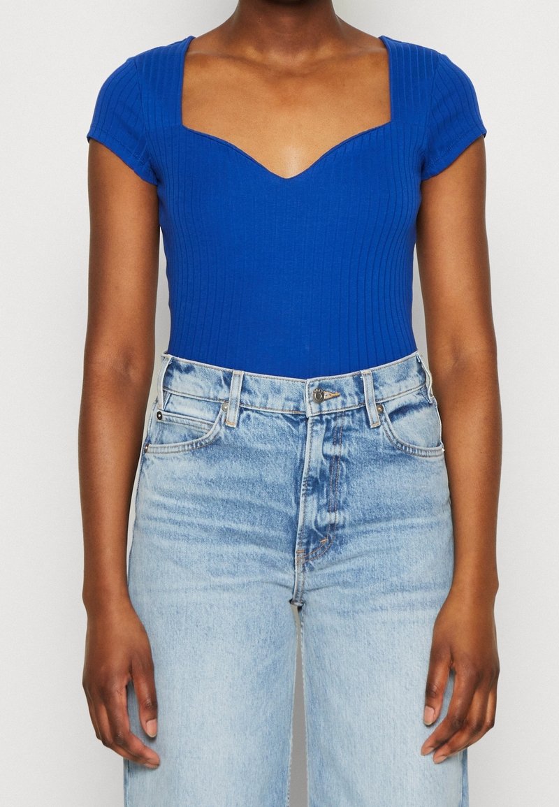 Personne portant un haut bleu côtelé à manches courtes avec un col carré et un jean taille haute bleu clair sur un fond uni.