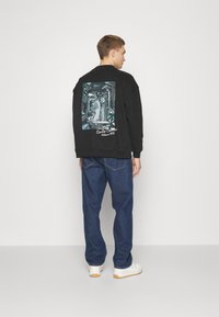 Zwart sweatshirt met een grafische print op de achterkant met een gestileerd gezicht. Gekleed met blauwe jeans en witte sneakers.