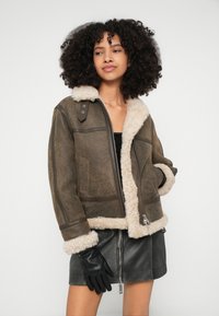 Hnedá bunda z umelého kože s krémovým kožušinovým golierom a manžetami; má bočné vrecká, predný zips a textúrovaný povrch.