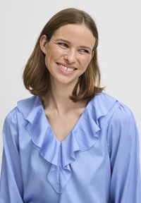 Blusa azzurro chiaro con scollatura a V, caratterizzata da un colletto arricciato e maniche lunghe. Il tessuto appare liscio e lucido con una leggera caduta.