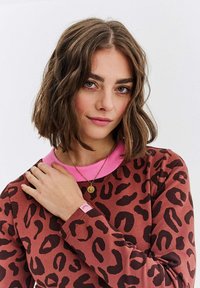 Mladá žena s krátkými vlnitými hnědými vlasy, oblečená v červeném topu s leopardím potiskem a růžovým límcem, s zlatým náhrdelníkem.