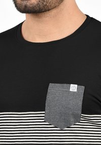 Camiseta negra de manga corta con rayas horizontales en blanco y negro, que presenta un bolsillo de un patrón gris oscuro y una pequeña etiqueta en el bolsillo.