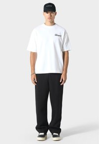 Biała koszulka z krótkim rękawem z małym czarnym napisem, zestawiona z luźnymi czarnymi spodniami i czapką w kolorze czarnym. Obuwie ma czarną cholewkę i białą podeszwę.