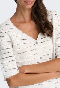 Femme portant un cardigan en tricot blanc à manches courtes avec des boutons décoratifs, bras croisés, montrant un bracelet de perles au poignet gauche.