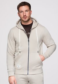 Homme portant un sweat à capuche beige zippé avec des cordons blancs et un pantalon assorti, debout, une main dans la poche, sur un fond uni.