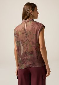 Blusa senza maniche in tessuto trasparente a fantasia floreale con colletto alto e chiusura a bottoni sul retro. I colori includono un mauve tenue e verde.