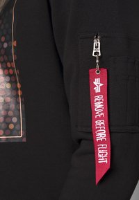 Sudadera negra con un diseño de puntos multicolores impresos, bolsillo con cremallera y tirador de marca, y una etiqueta roja que dice "RETIRE ANTES DEL VUELO".