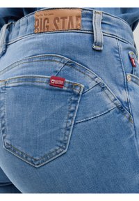 Valge taustaga hele siniste teksade tagantvaade, nähtavad õmblused, pruun nahast brändiplaat ja punane kangasildid tagataskul.