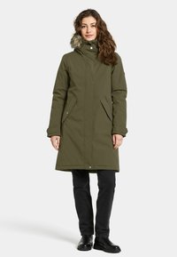Olijfgroen lange jas met een bontgevoerd capuchon, ritszakken en een rechte snit. Zwarte broek en laarzen maken de outfit compleet.