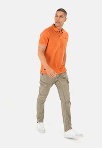 Oranje polohemd met een klassieke kraag, gecombineerd met lichtbeige cargo-broeken met meerdere zakken en een relaxte pasvorm, gedragen met witte sneakers.