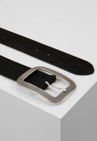 Cinturón de ante negro con una hebilla cuadrada de plata texturizada. Superficie lisa con múltiples agujeros de ajuste a lo largo de la correa.
