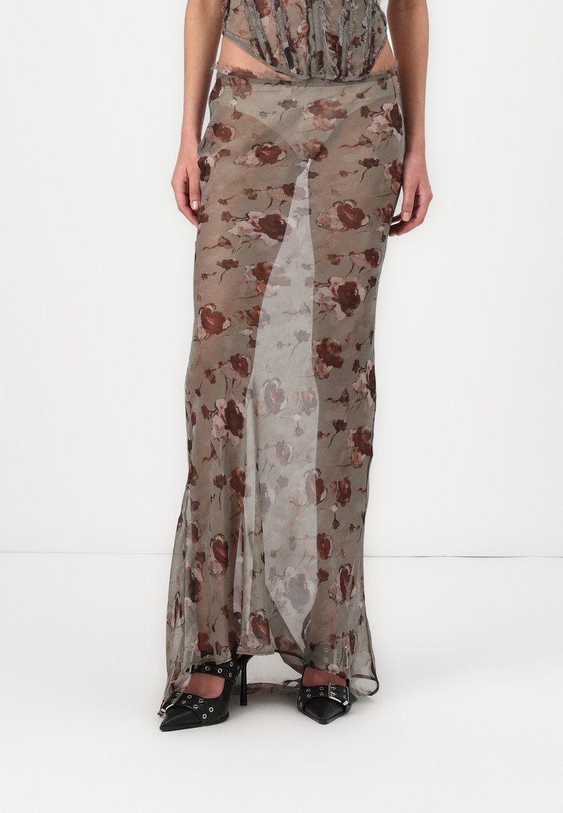 Maxi gonna floreale trasparente in verde tenue, con un motivo di rose marroni, spacchi alti e un design fluido. Indossata con tacchi neri a punta.