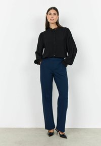 Sort bluse med blonderkrave og lange ærmer, parret med navy bukser og sorte hæle, poserende mod en ensfarvet hvid baggrund.
