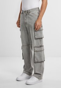 Pantaloni cargo grigi in denim, con multiple tasche laterali, vestibilità ampia e cuciture a vista. Abbinati a sneakers bianche.