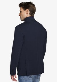 Mørkeblå blazer med en glat tekstur, slagkrave og sidelommer. Har knappede ærmer og en skræddersyet pasform. Placeret på en mands ryg.