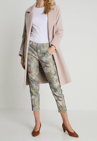 Manteau en laine rose pâle sur un t-shirt blanc, associé à un pantalon ajusté à motif floral en vert et marron. Des talons en suède marron complètent le look.