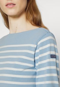 Pull bleu clair à rayures en tissu tricoté. Présente des rayures blanches horizontales et un petit écusson logo sur l'épaule gauche.
