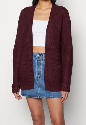 Vrouw draagt een kastanjebruine gebreide cardigan over een wit cropped topje en een blauwe spijkerrok, staand tegen een effen achtergrond.