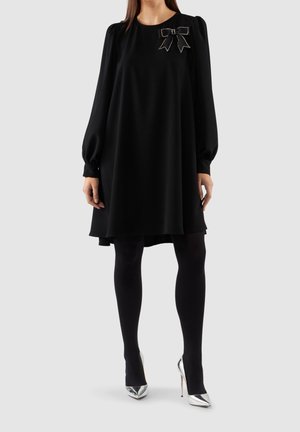 Femme portant une robe noire à manches longues avec un nœud décoratif sur la poitrine, des collants noirs et des chaussures à talons hauts argentées.