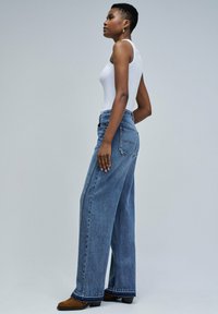 Woman in white sleeveless top, high-waisted wide-leg blue jeans, and brown pointed shoes, standing sideways against a plain background.