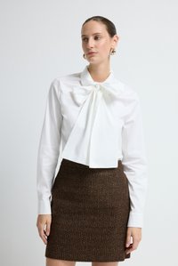Witte blouse met lange mouwen en een grote strik bij de kraag, gecombineerd met een nauwsluitende bruine rok met een textuurpatroon.