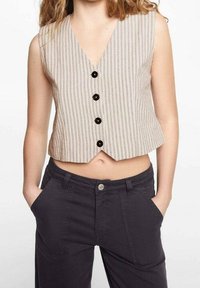 Béžové pruhované tílko bez rukávů s výstřihem do V a pěti černými knoflíky vpředu; nošené přes tmavé, těsné kalhoty.