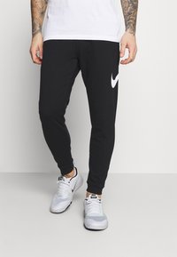 Pantalons de survêtement noirs ajustés avec des poignets élastiques, présentant un grand logo Nike blanc sur la jambe gauche. Portés avec des baskets blanches.