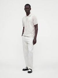 Homme portant un polo à manches courtes à motif clair, un pantalon blanc et des mocassins noirs et blancs, debout, les mains dans les poches sur fond blanc.
